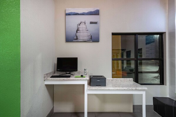 A workspace with a desk, computer, and printer, a window with a view of a building, and a painting of a dock and mountains on the wall.