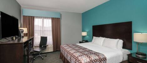 A hotel room with a large bed, a desk with a TV, a chair, and a nightstand with a lamp.