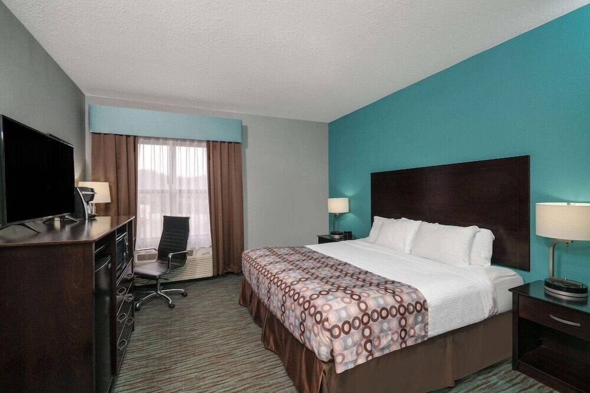 Una camera d'albergo con un letto grande, una scrivania con televisione, una sedia, una finestra con tende e un comodino con una lampada.