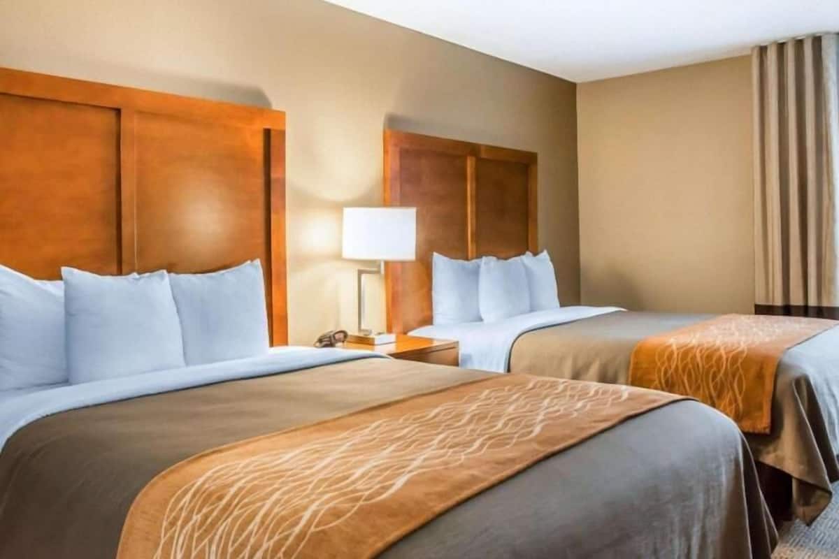 A hotel room with two beds, wooden headboards, a nightstand with a lamp, and a window with curtains.