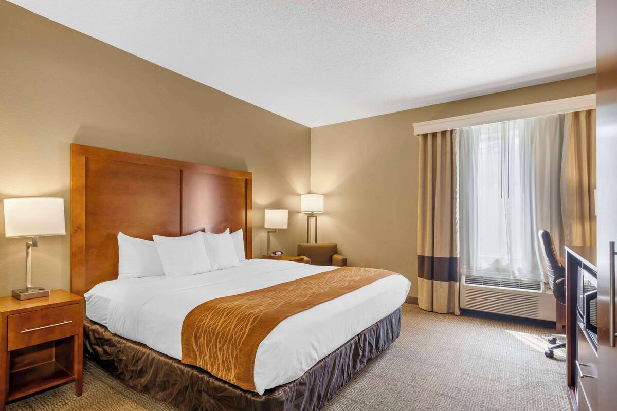 A hotel room with a large bed, two bedside tables with lamps, a chair, a TV, and a window with curtains.