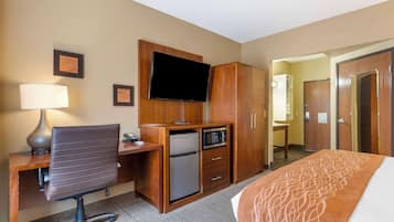 A hotel room with a bed, desk, chair, television, wardrobe, and a small refrigerator.