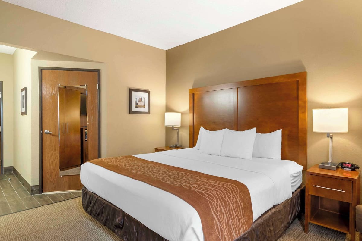 A hotel room with a large bed, wooden headboard, two bedside tables with lamps, a nightstand, and a door.