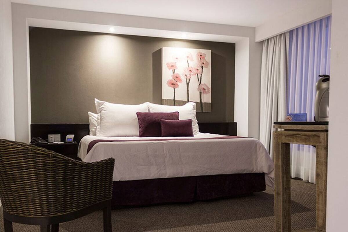 A hotel room with a bed, a chair, a nightstand, a lamp, and a painting on the wall.