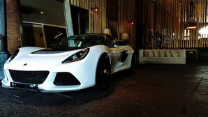 Um carro desportivo conversível branco estacionado numa área interior bem iluminada, com um sofá e elementos decorativos ao fundo.