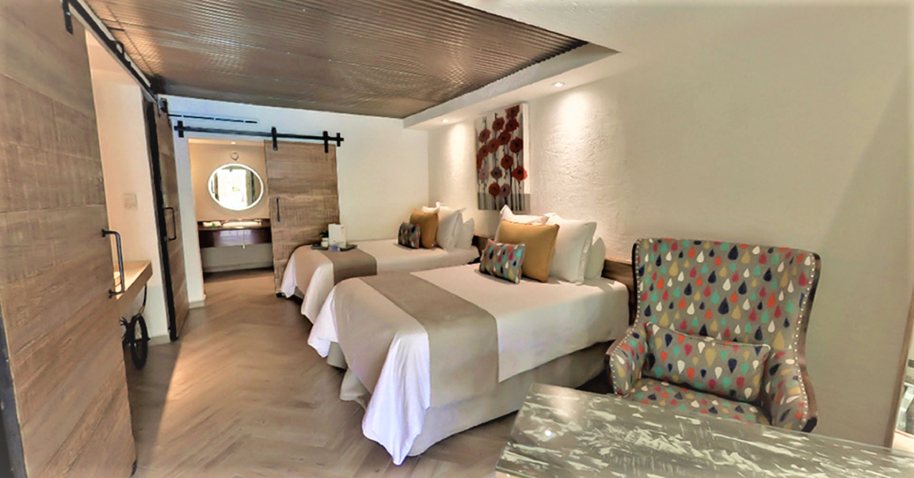 A hotel room with two beds, a chair, a table, a mirror, and a bathroom with a sink and mirror.
