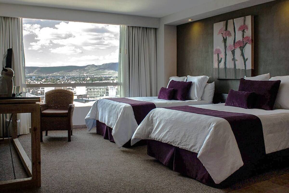 A hotel room with two beds, a desk, a chair, and a large window with a view of a cityscape.