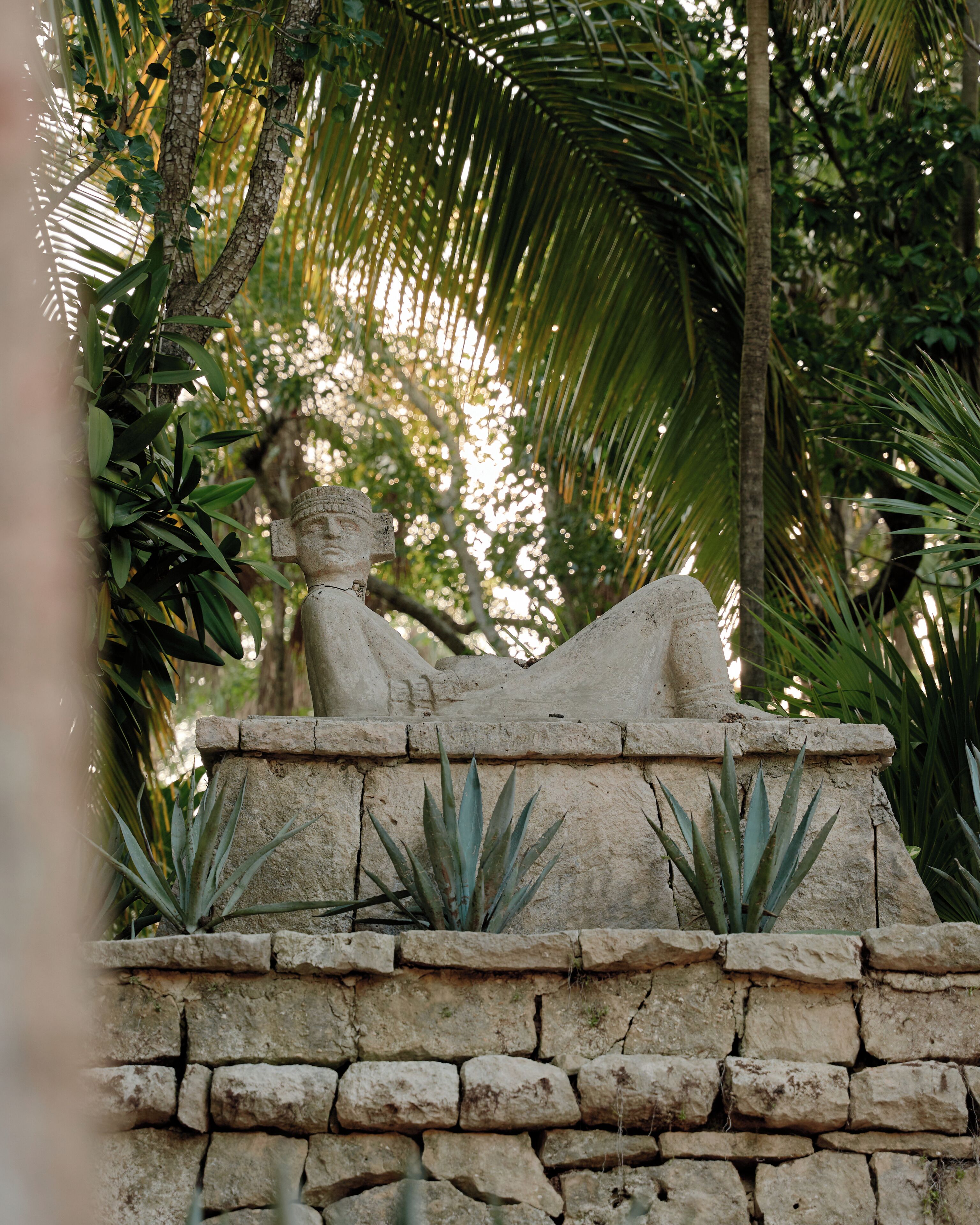 A stone statue of a reclining figure on a stone platform with plants.