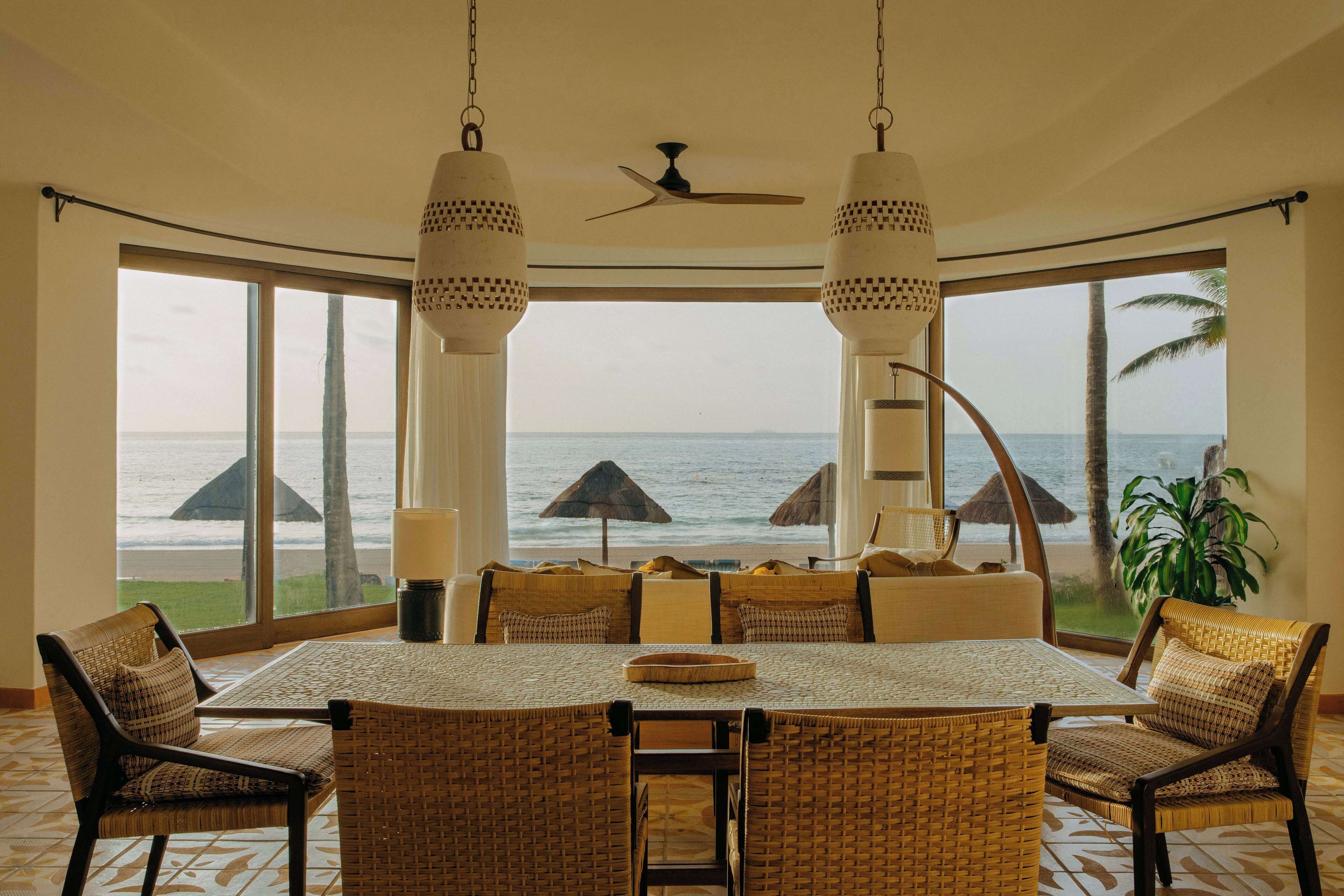 A dining area with a view of the ocean, featuring a large table, wicker chairs, and hanging lamps.