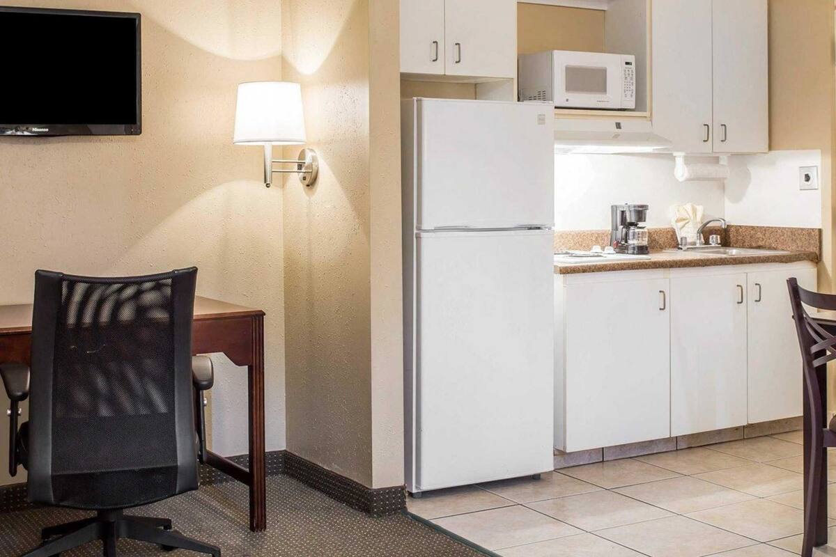A compact hotel kitchen with white cabinets, a microwave, a refrigerator, a coffee maker, and a dining area with a chair and table.