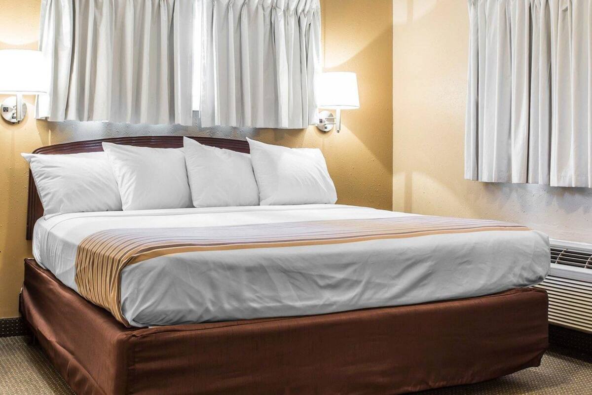 A hotel room with a bed, two bedside lamps, a window with curtains, and a wall-mounted air conditioning unit.