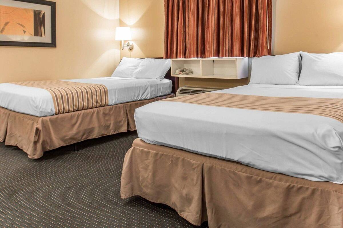 A hotel room with two beds, a nightstand, a lamp, a painting on the wall, and a window with curtains.
