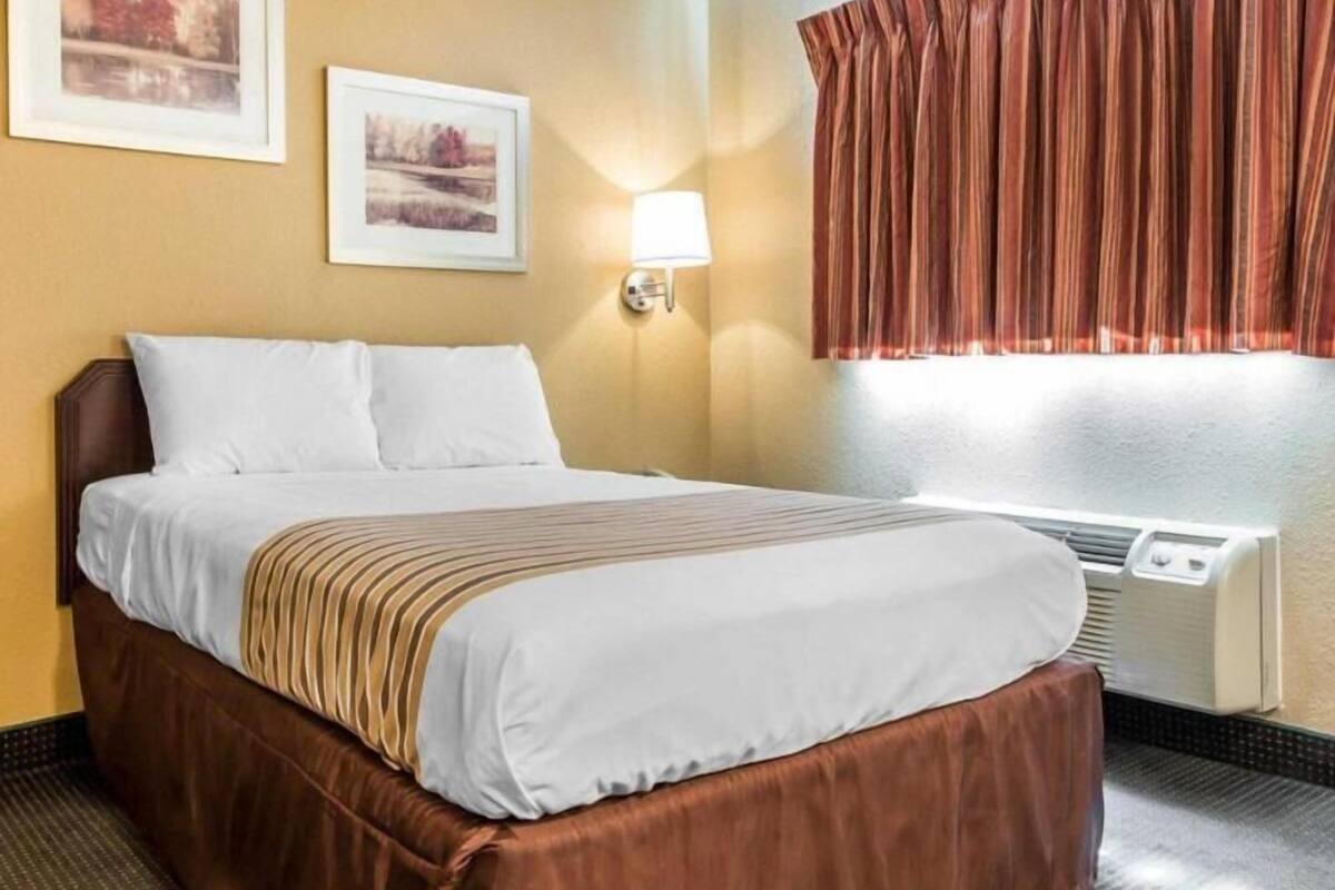 A hotel room with a bed, two framed pictures on the wall, a window with curtains, and a wall-mounted lamp.