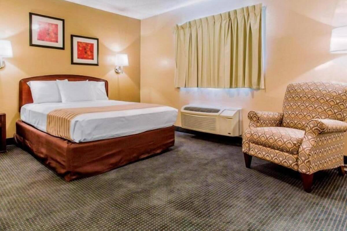 A hotel room with a bed, a chair, a wall-mounted air conditioner, and two framed pictures.