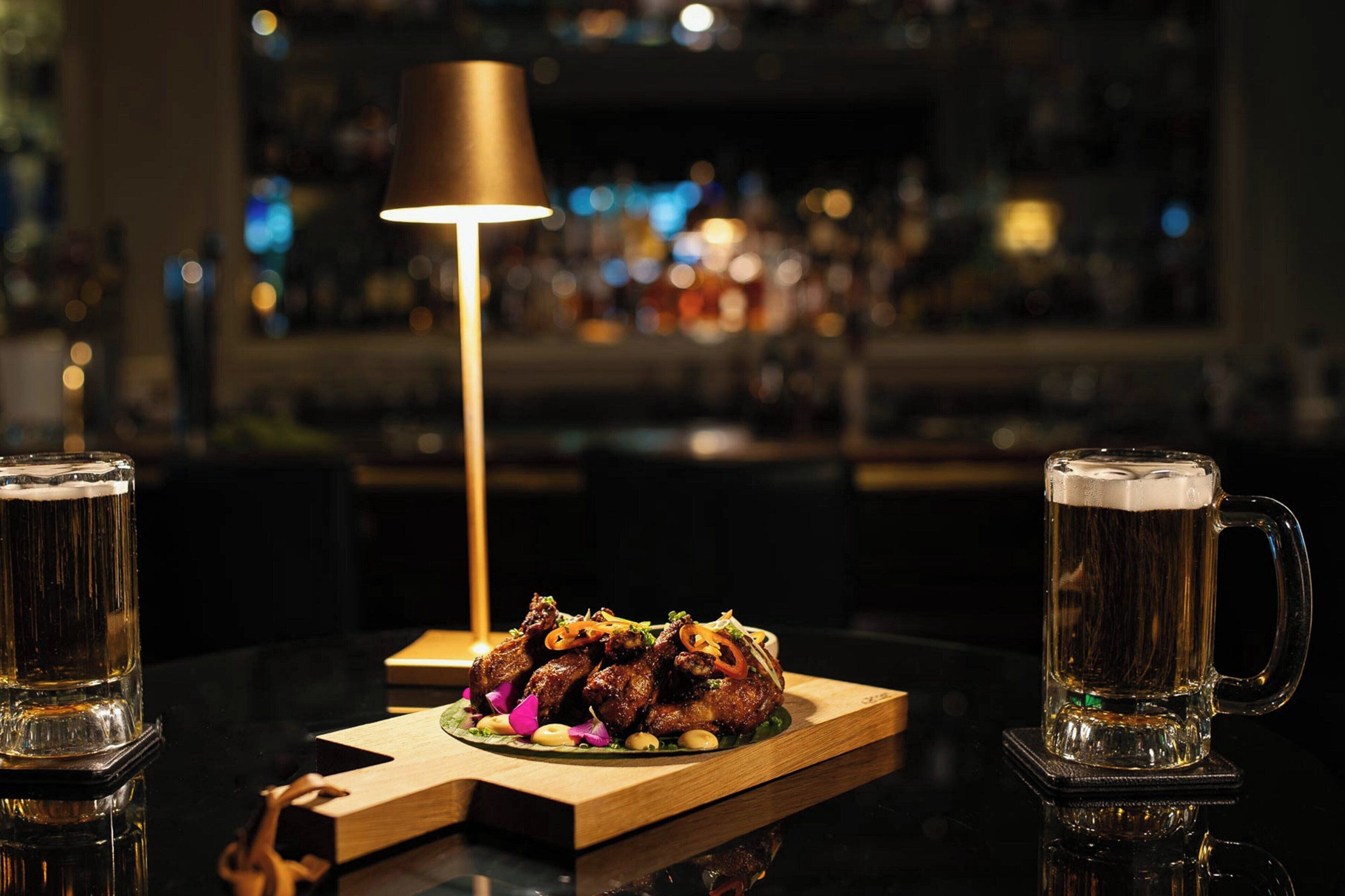 Une scène de bar avec une lampe, une assiette de nourriture et deux chopes de bière.