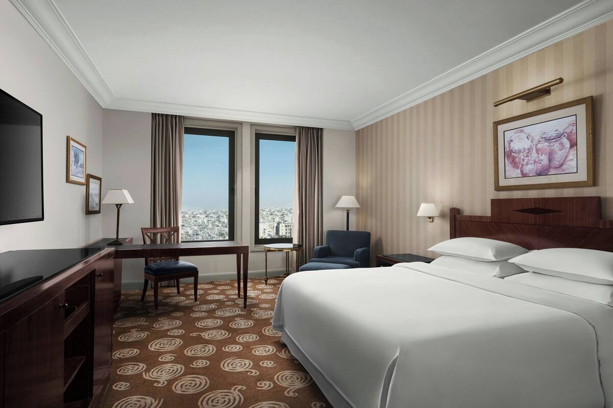 A hotel room with a large bed, a desk, a chair, two windows, and a television.