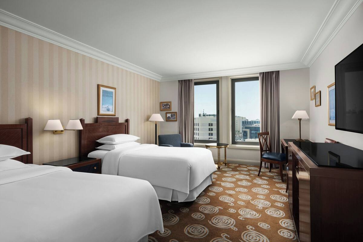 A hotel room with two beds, a desk, a chair, a TV, and a window with a city view.