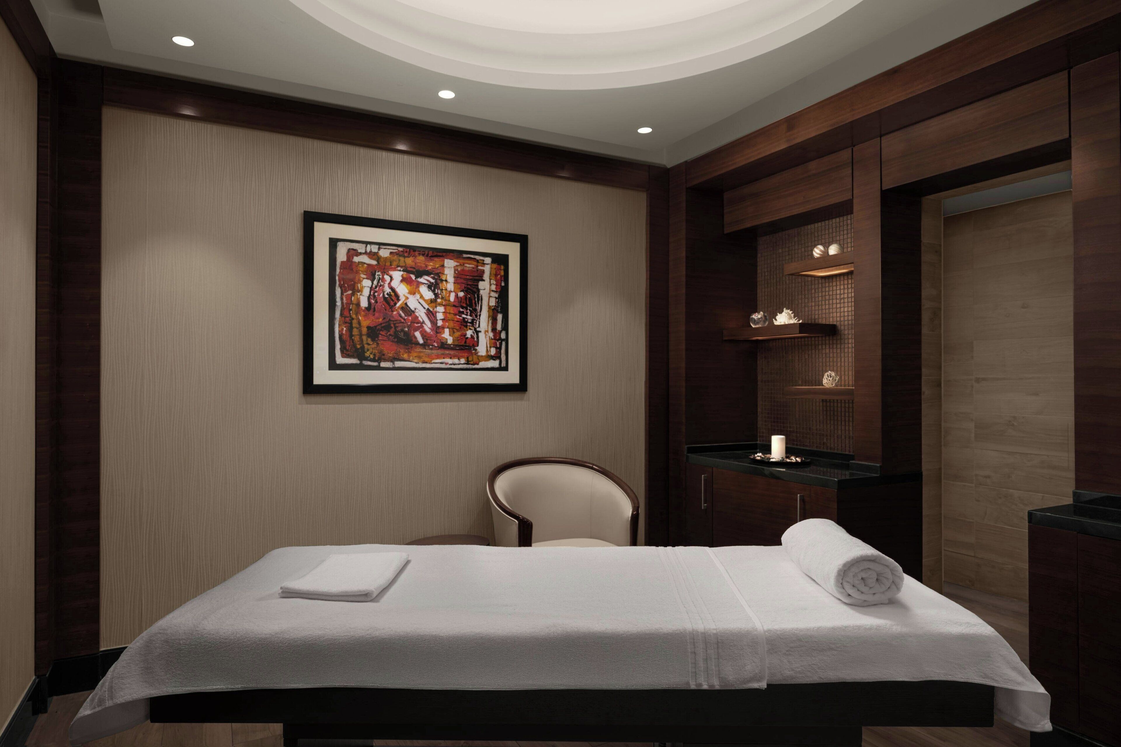 A spa room with a massage table, a chair, a framed abstract painting, and a shelf with candles.