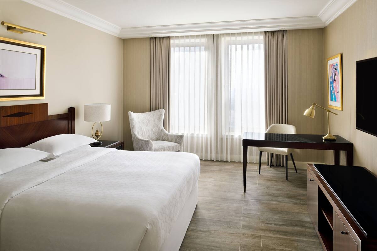 A hotel room with a large bed, a desk with a lamp, a chair, and a TV.