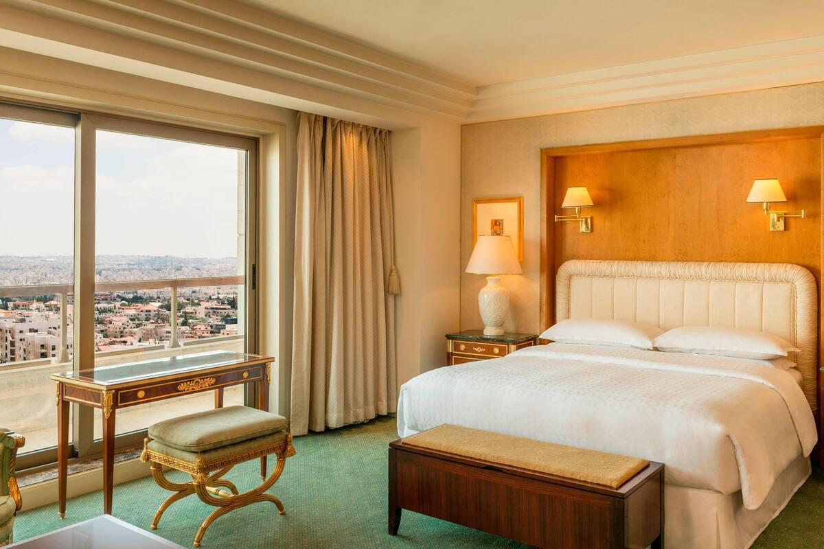 A hotel room with a large window, a bed, a bedside table with a lamp, and a small bench.