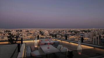 A rooftop terrace with a dining area, seating, and a city view at dusk.
