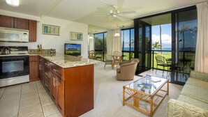 A modern kitchen with a large island, a dining area with chairs, and a view of the ocean through large sliding glass doors. - Kihei Surfside - Maui Condo & Home (Kihei)