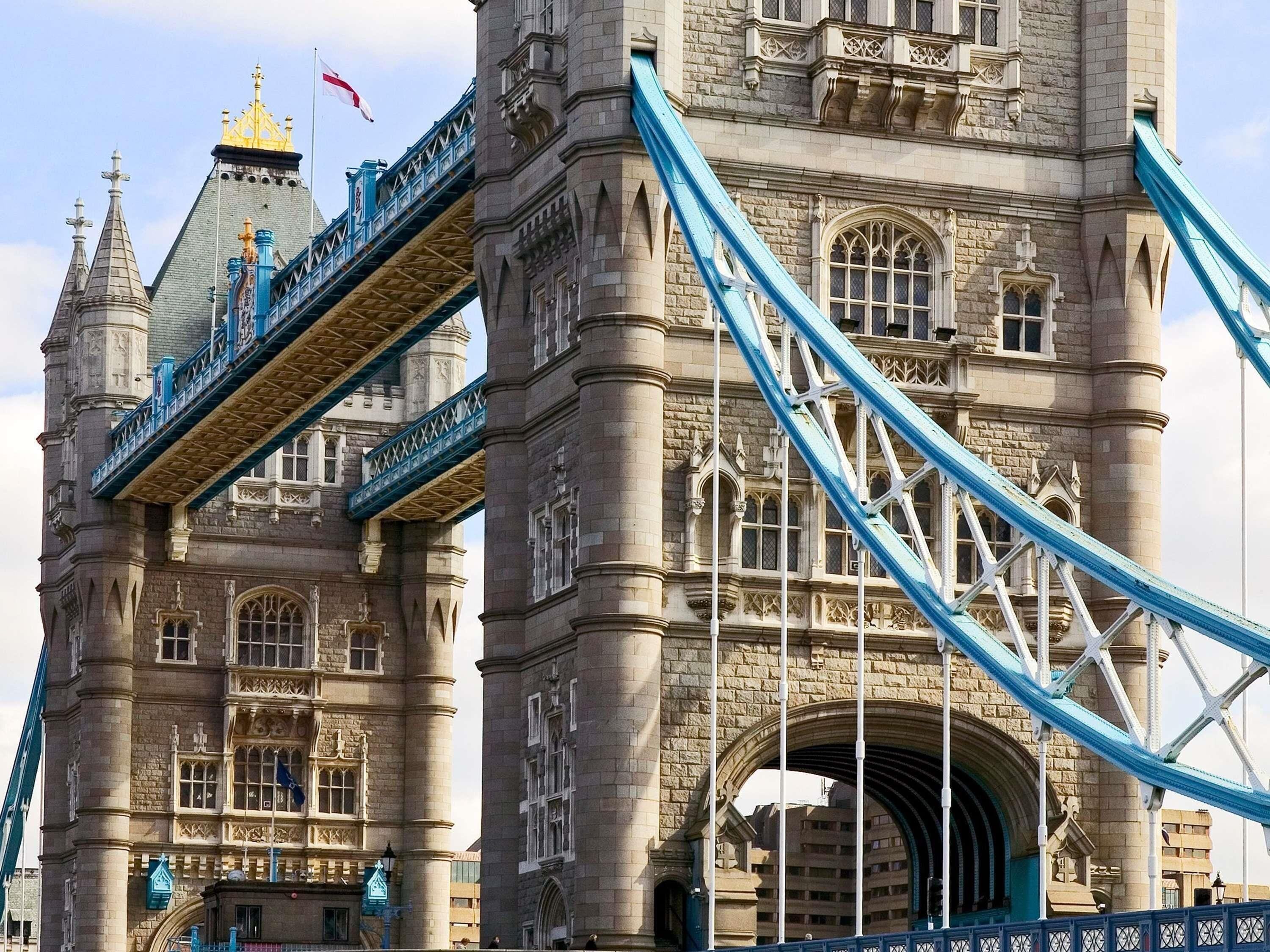 A historic stone tower with a large archway, a bridge with blue railings, and a flag on top.