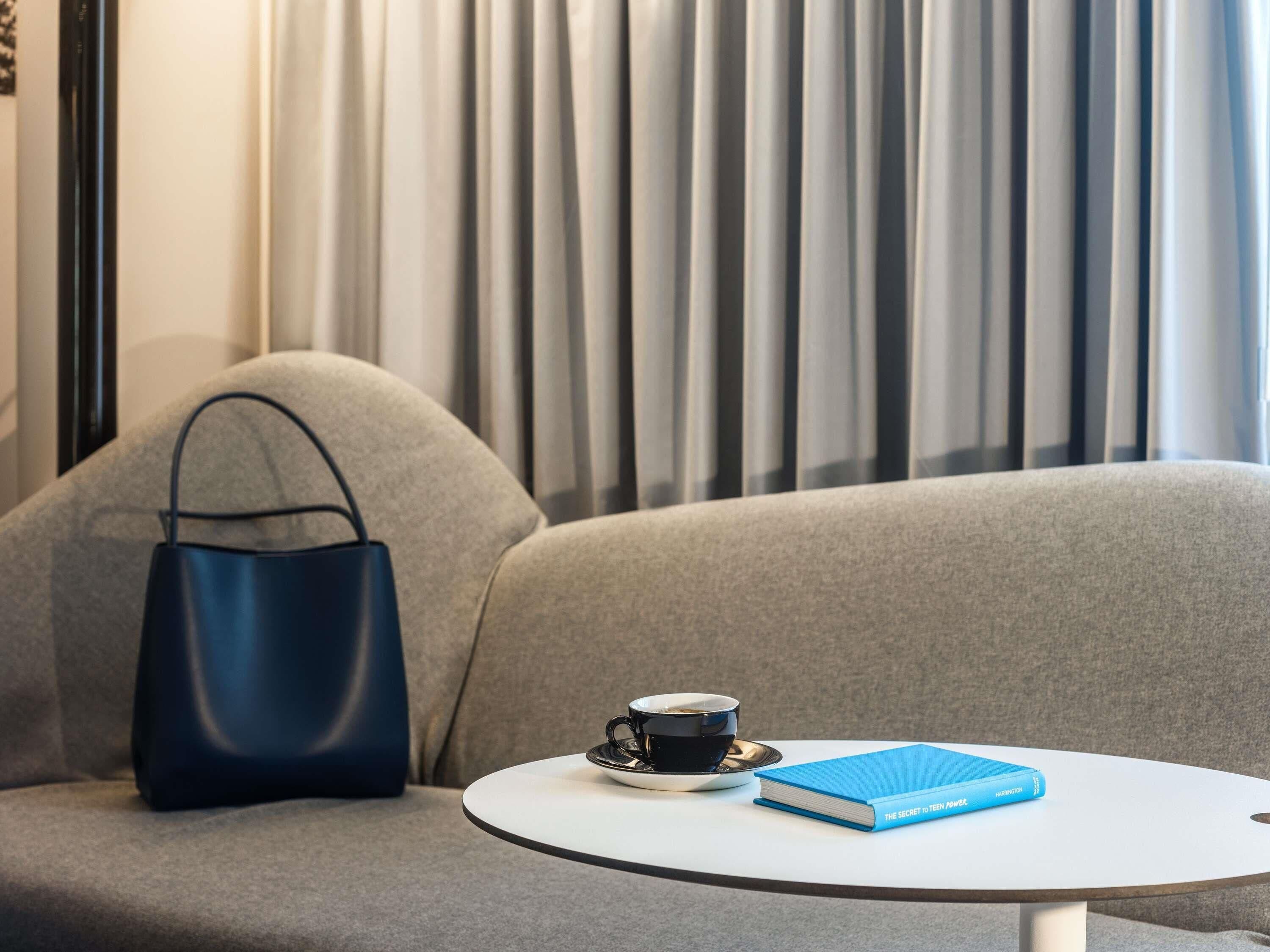 A modern room with a grey sofa, a blue handbag, a white table with a book and a cup of coffee.