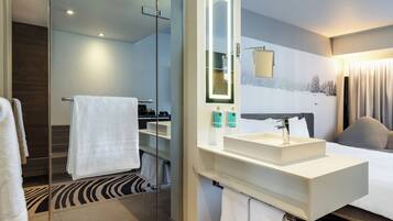 Une chambre d’hôtel moderne dotée d’une douche en verre, d’un grand miroir, d’un lavabo blanc et d’un lit avec une tête de lit.