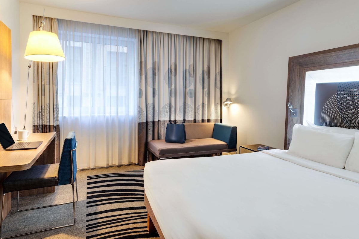 Hotellihuone, jossa on sänky, työpöytä kannettavalla tietokoneella, tuoli, sohva ja lamppu.