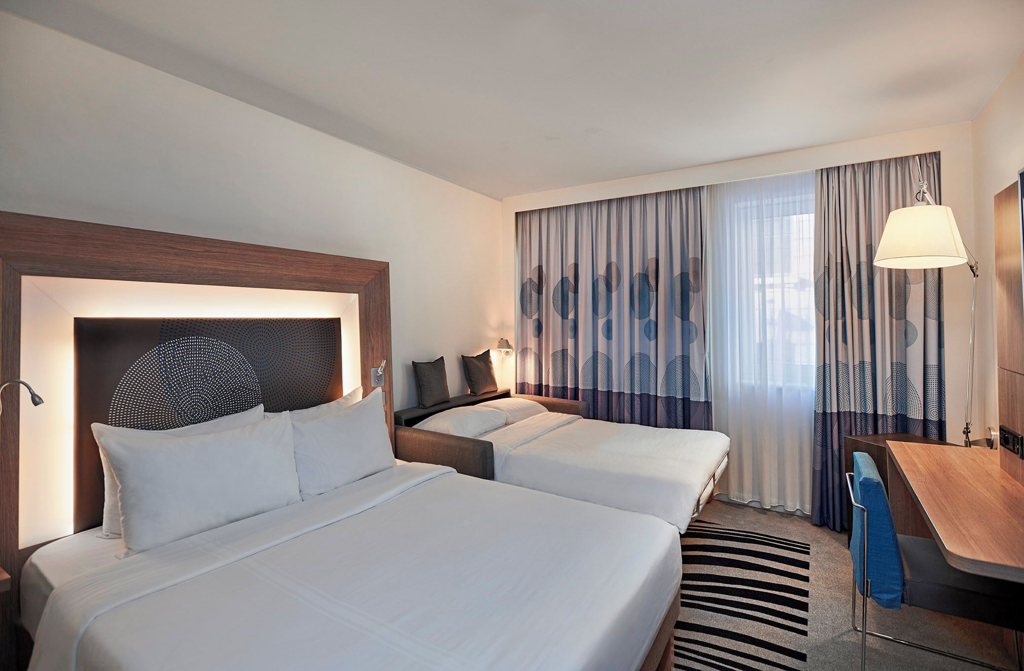 A hotel room with two beds, a desk, a lamp, and a view of the outdoors through curtains.