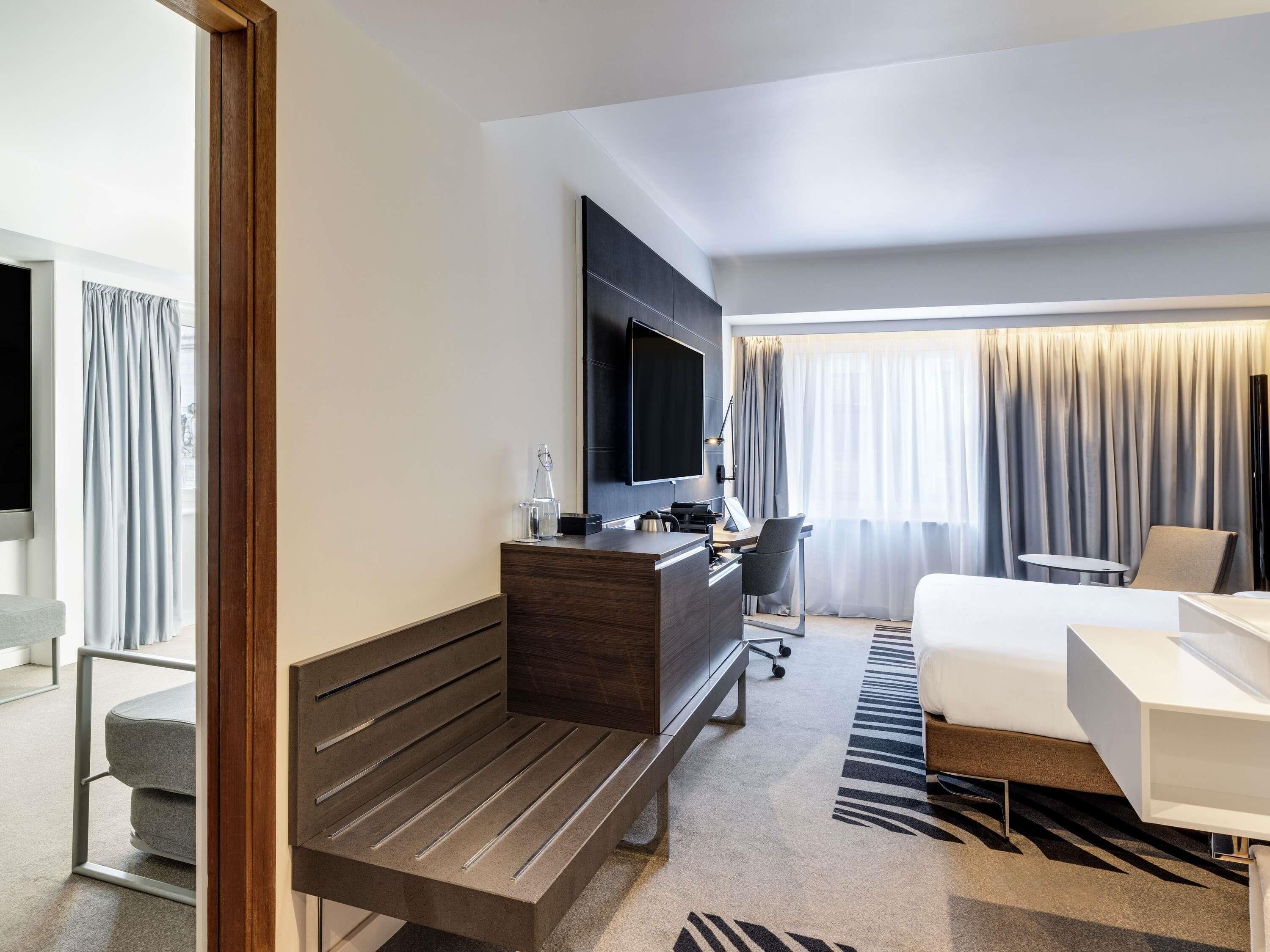 A modern hotel room with a flat-screen TV, a desk with a chair, a sofa, a bench, and a patterned rug.