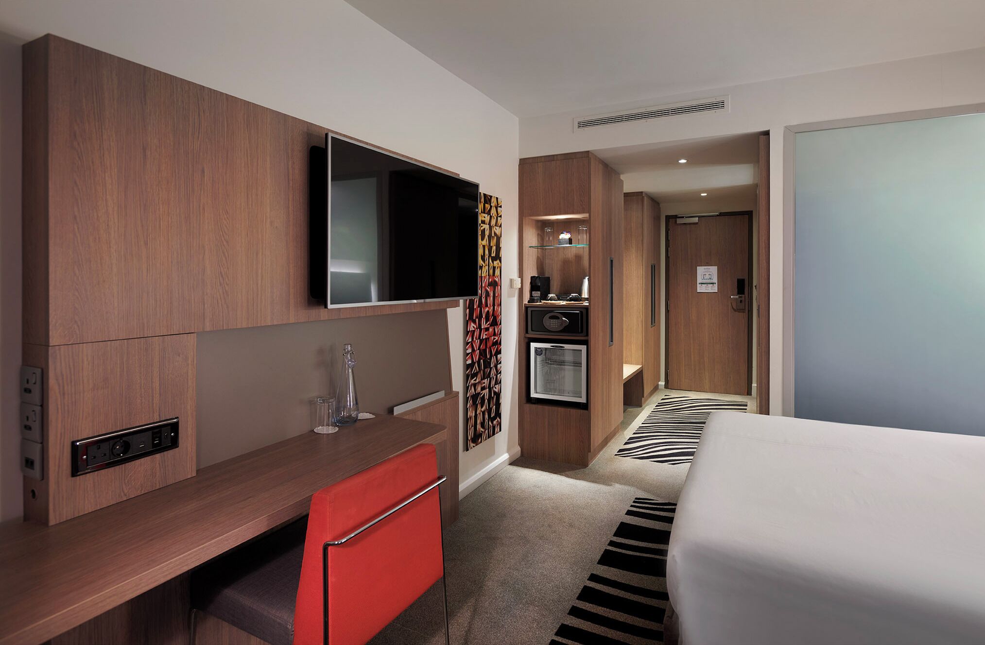 Ett hotellrum med en säng, ett skrivbord med en stol, en tv, en minibar och en dörr som leder till ett annat rum.