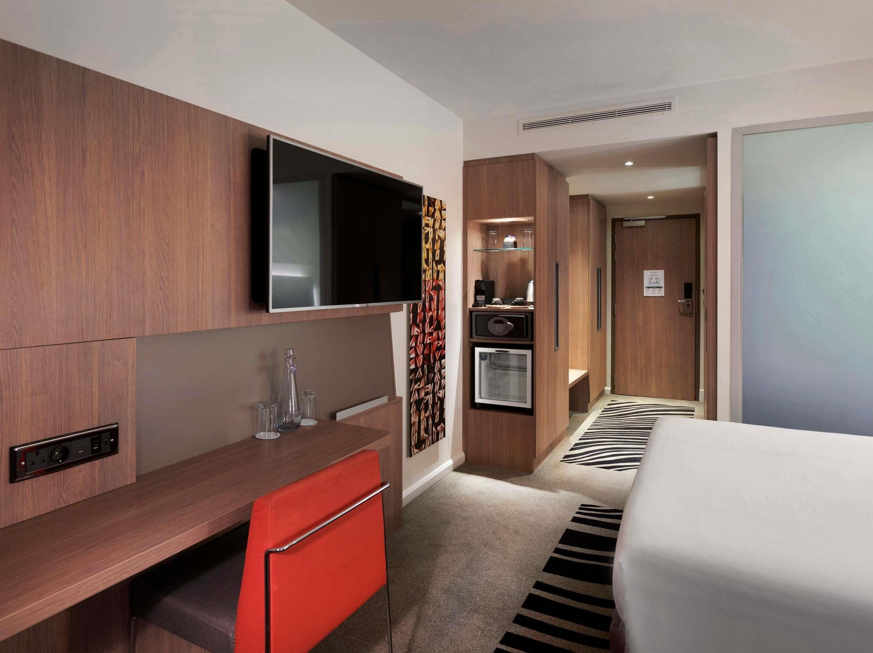 Quarto de hotel com cama, mesa com cadeira, televisão, minibar e porta que dá para outro aposento.