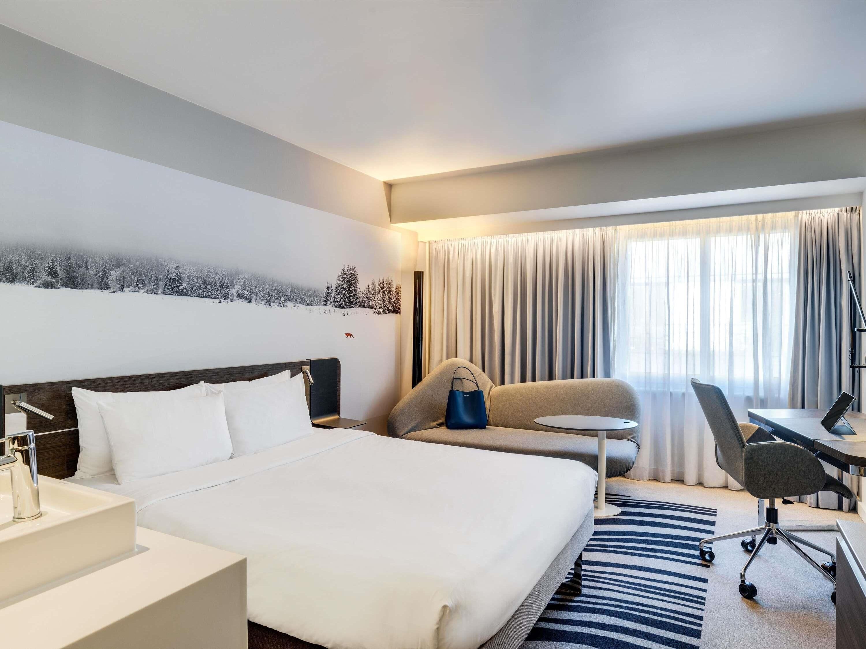 Quarto de hotel com cama, escrivaninha, cadeira, mesinha de centro e uma grande janela com cortinas.