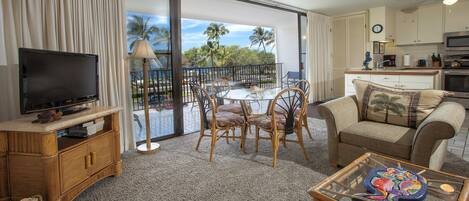 Un salon avec un téléviseur, une table à manger avec des chaises, un canapé et un balcon offrant une vue sur les palmiers.
