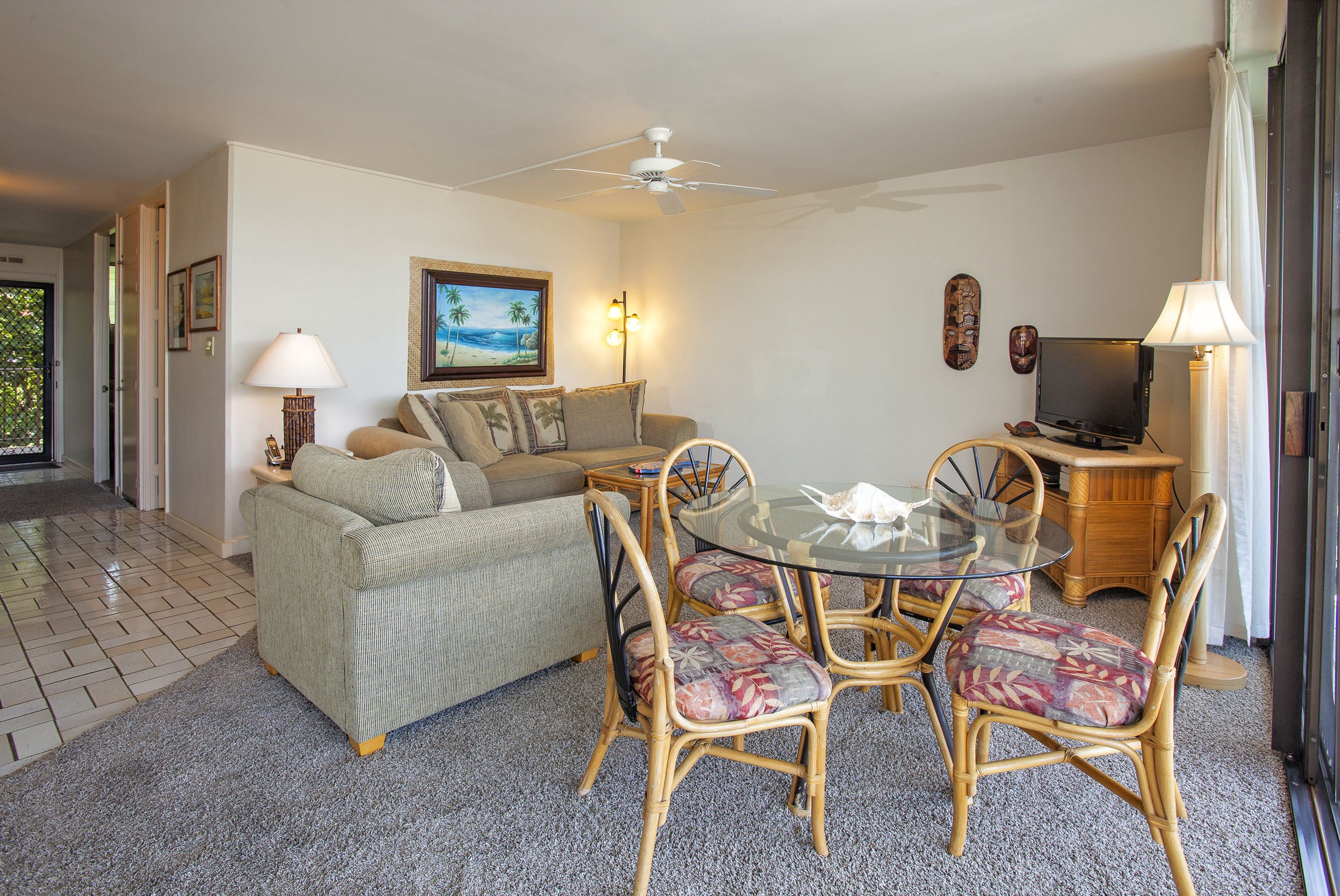 A living room with a sofa, a glass-top dining table with chairs, a ceiling fan, and a television.