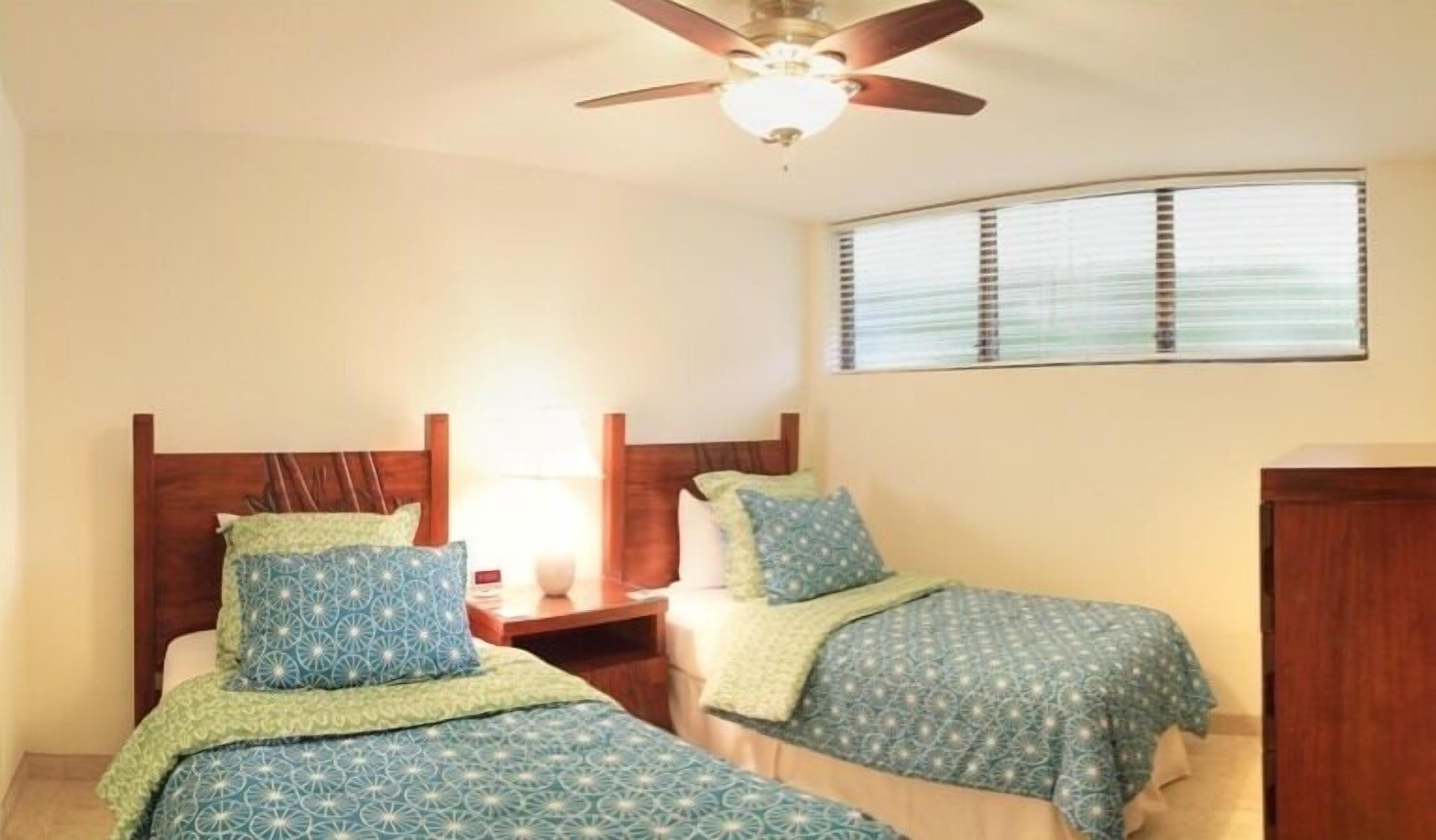 A bedroom with two beds, a ceiling fan, a window with blinds, and a nightstand.