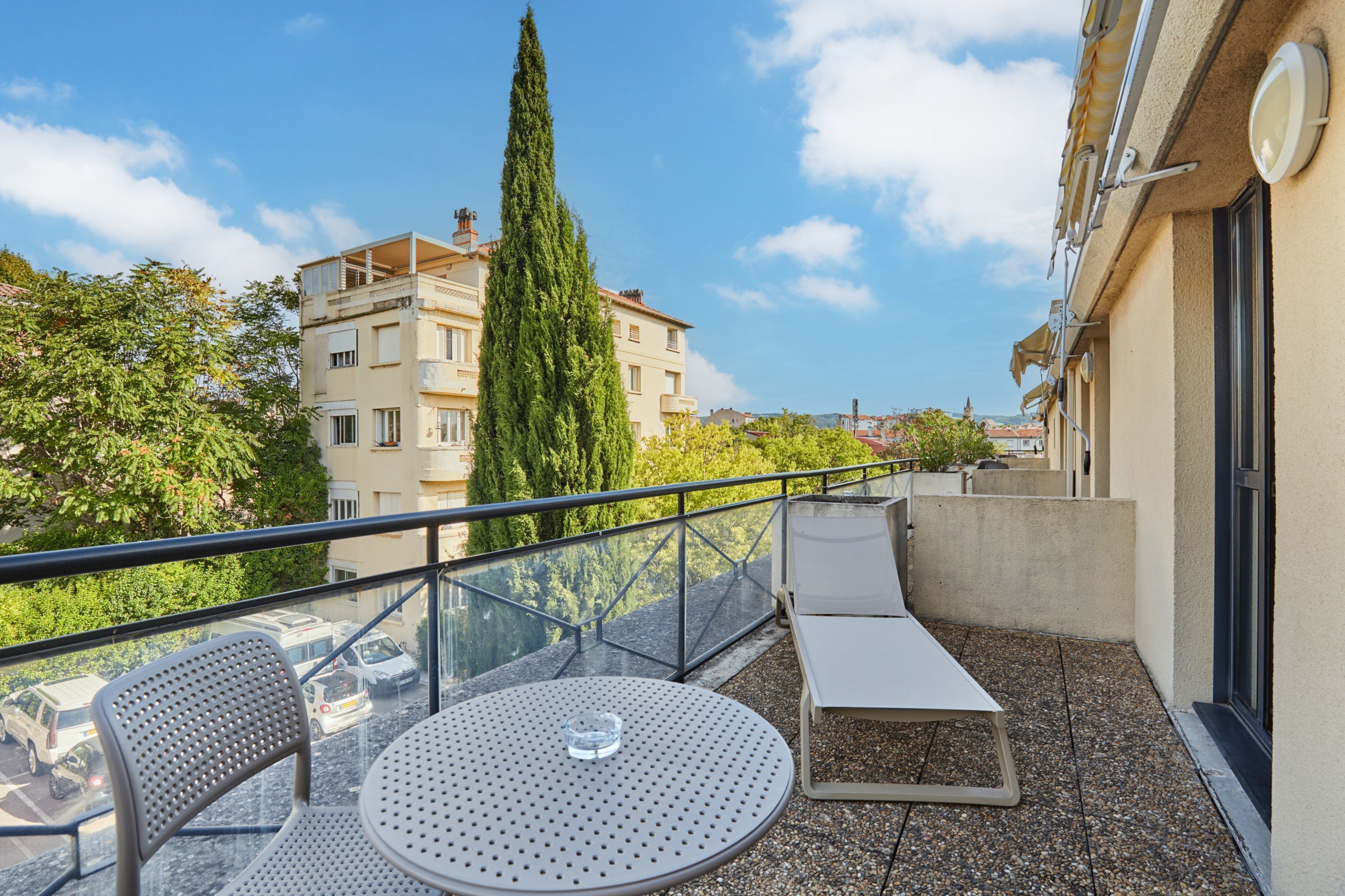 Ein Balkon mit rundem Tisch, zwei Stühlen, einer Liege und einem hohen Baum.