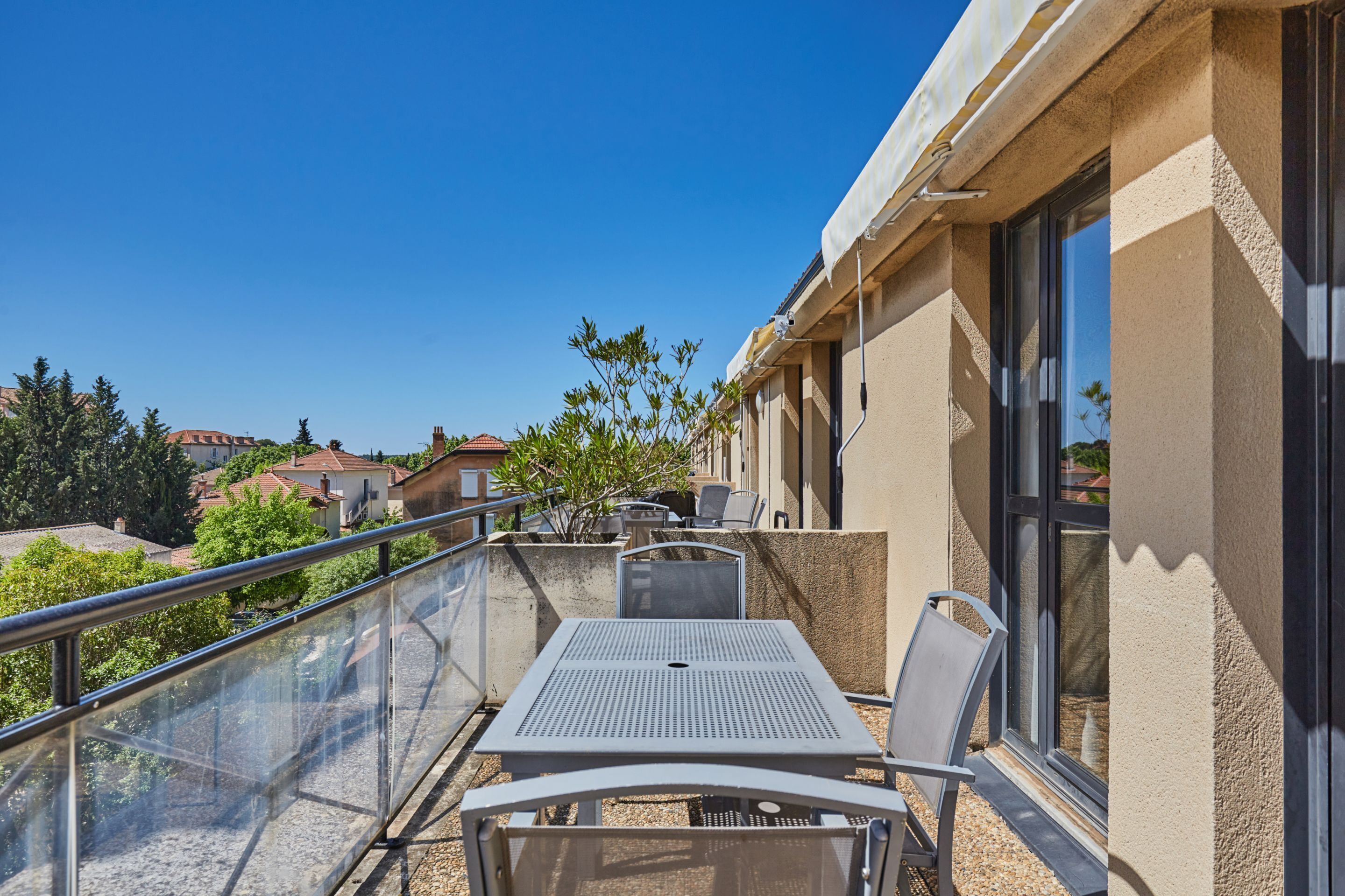 En balkon med et bord og stole, gelænder af glas og udsigt til omkringliggende bygninger og træer.