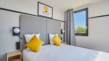 A neatly made bed with white and yellow pillows, a headboard, bedside tables, a wall-mounted lamp, and a window with curtains.