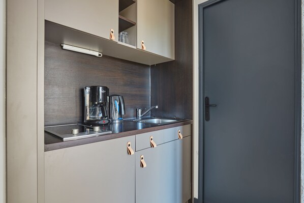 A compact kitchen with a coffee maker, kettle, and a small shelf with glassware.