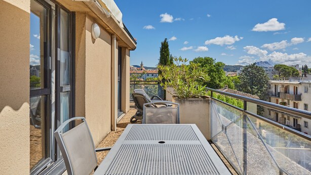 Un balcón con mesa y sillas, con vista a un paisaje urbano con edificios y una montaña al fondo.