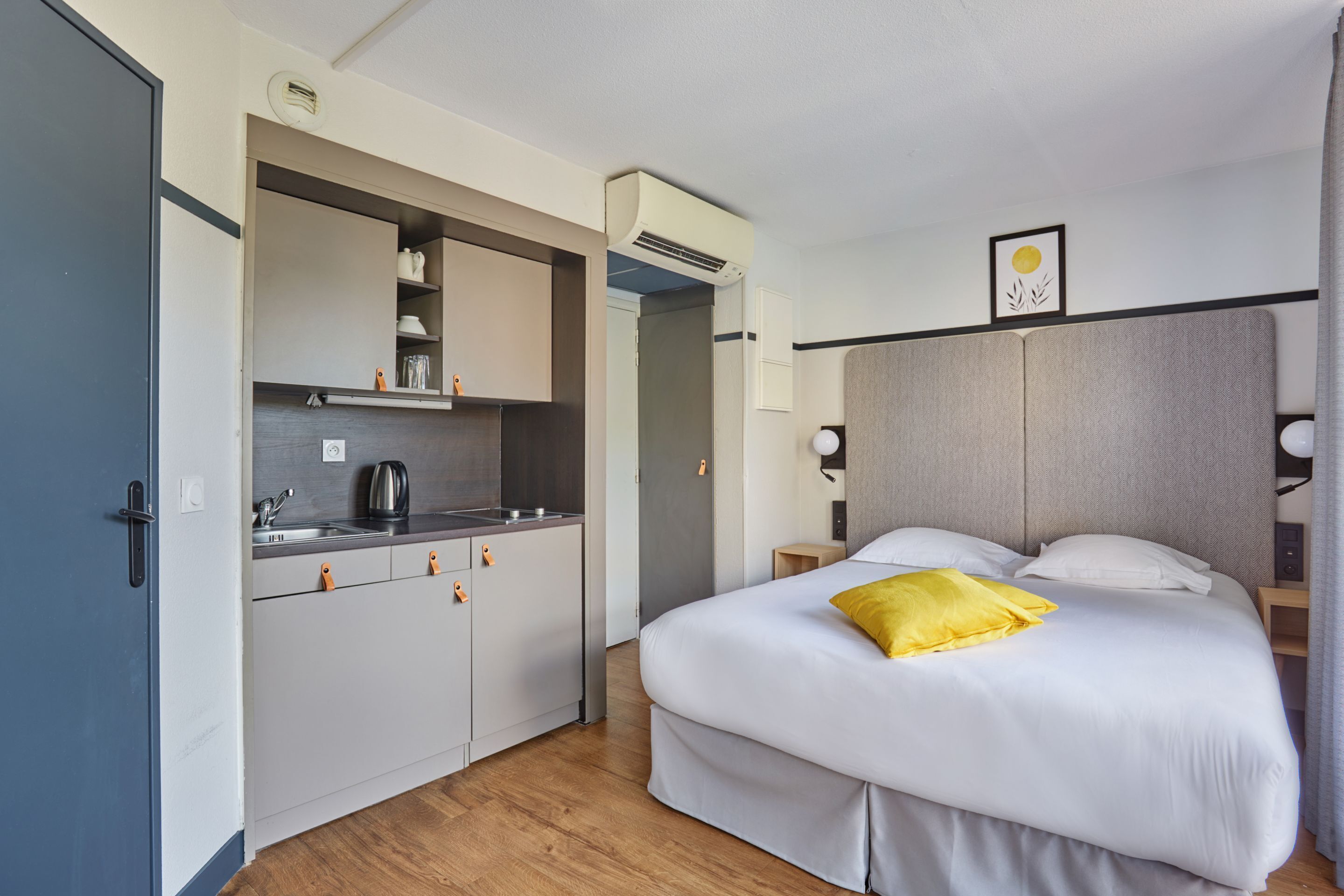 A compact hotel room with a kitchenette, a bed with white bedding and a yellow pillow, a bedside table with a lamp, and a framed picture on the wall.