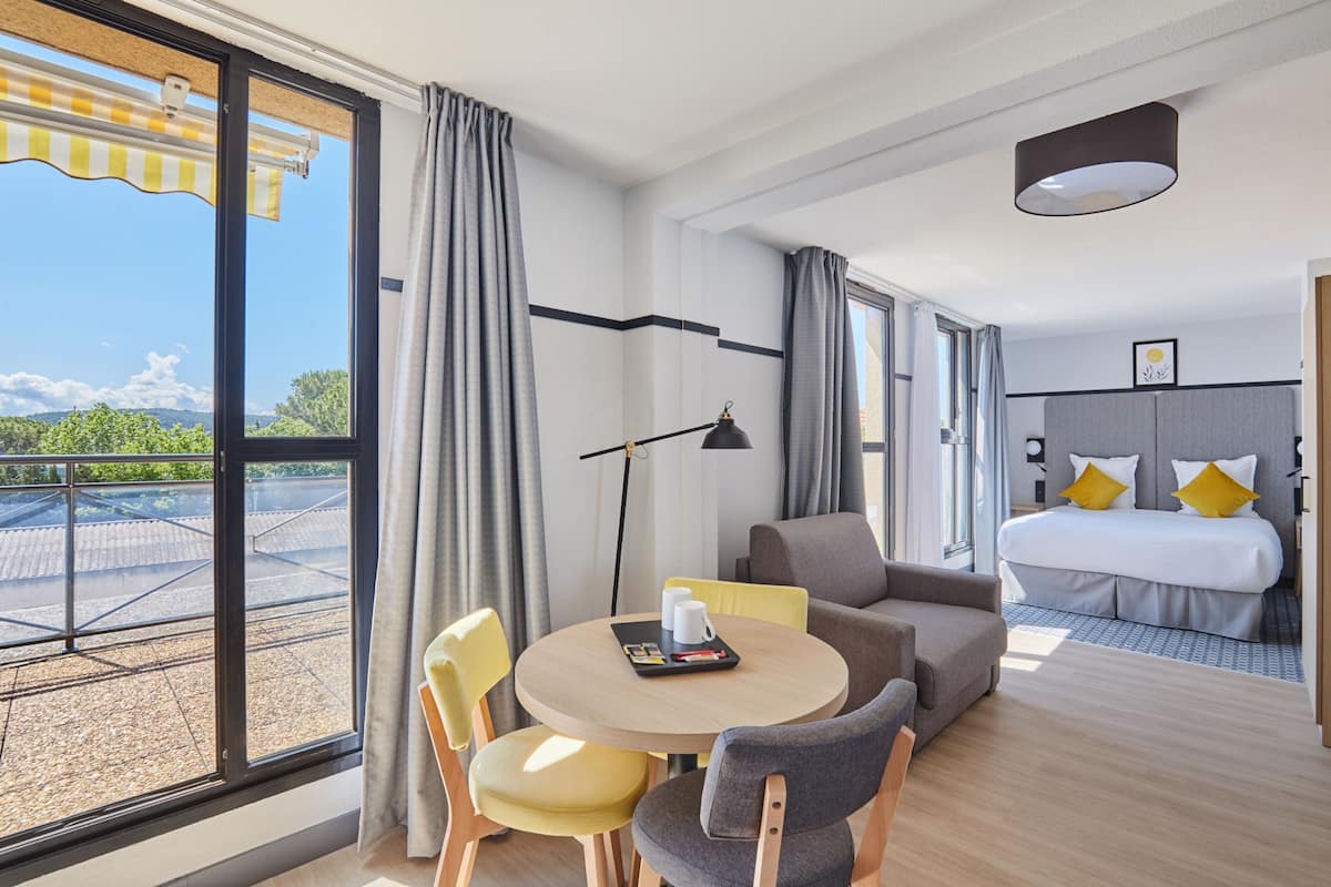 A modern hotel room with a balcony, a round dining table, a sofa, and a bed with a view of the outdoors.