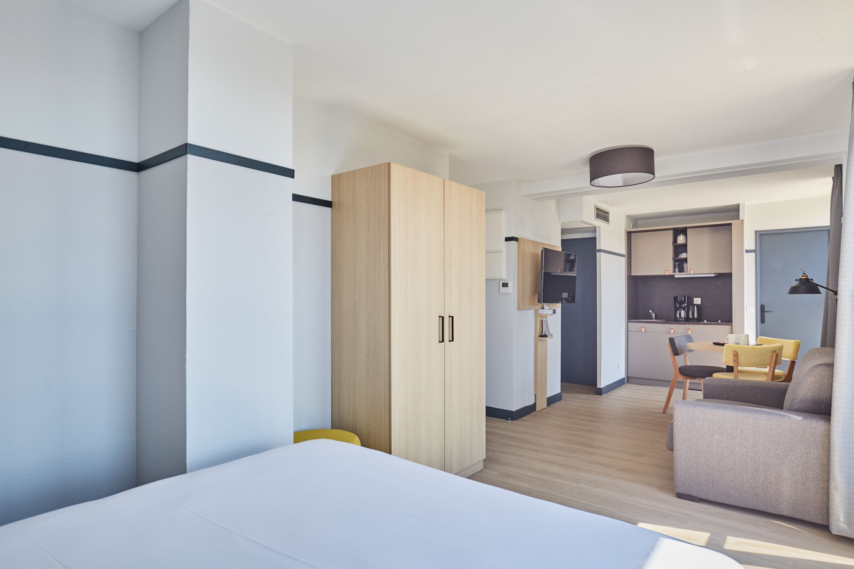 A modern hotel room with a bed, a wardrobe, a kitchenette, and a seating area.