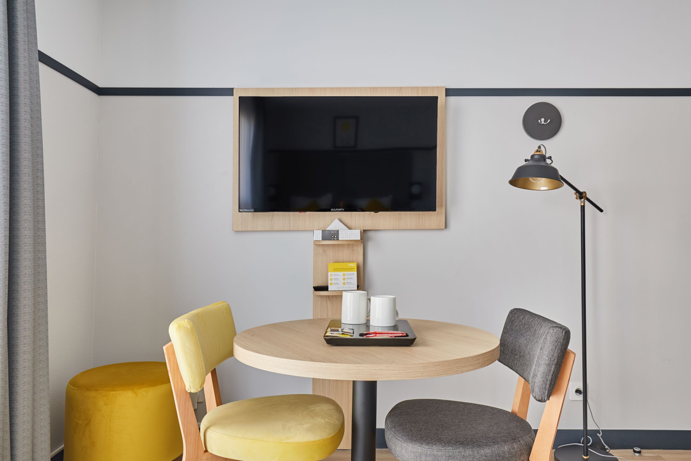 A modern room with a round wooden table, two chairs (one yellow, one grey), a mounted television, a floor lamp, and a small shelf with a box and two white mugs.