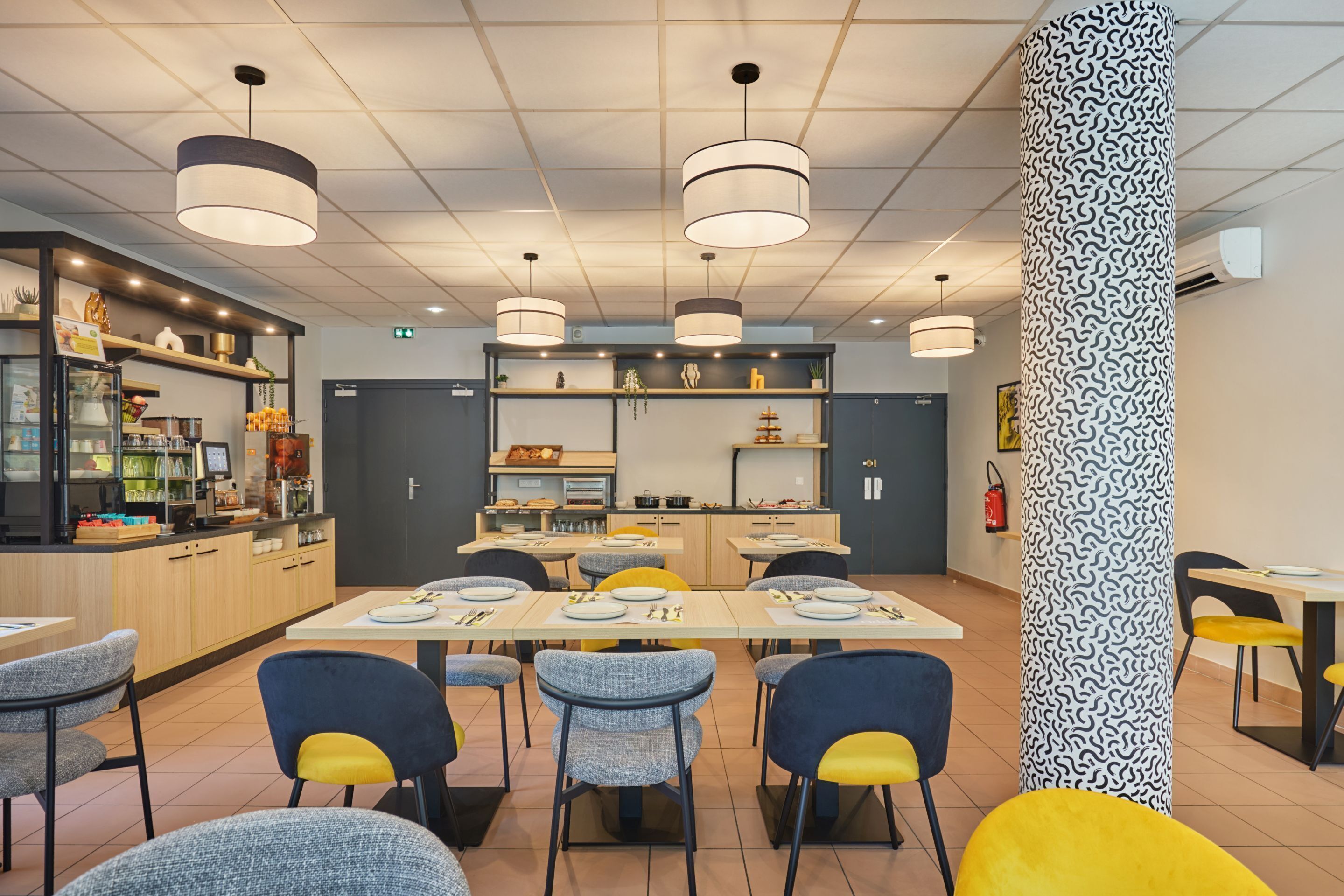 A modern dining area with multiple tables and chairs, a kitchen in the background, and a patterned column.