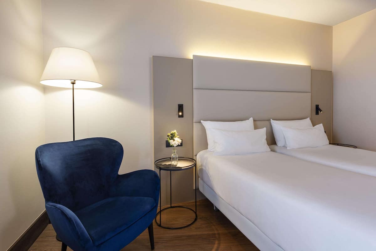 Una habitación de hotel con una cama grande, un sillón azul, una mesita redonda con un jarrón de flores y una lámpara de pared.
