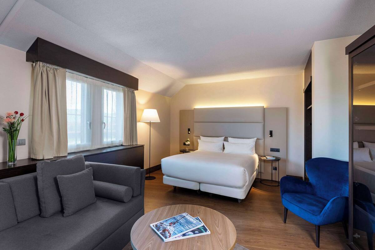 Una habitación de hotel moderna con una cama, un sofá, un sillón azul, una mesita de centro redonda y una mesita de noche con una lámpara.